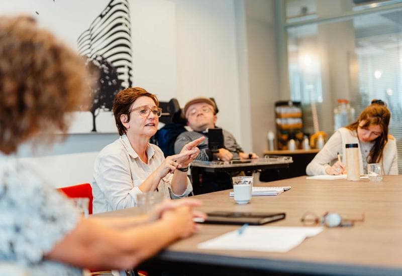 Vrouw zit aan tafel en legt iets uit aan een groep.