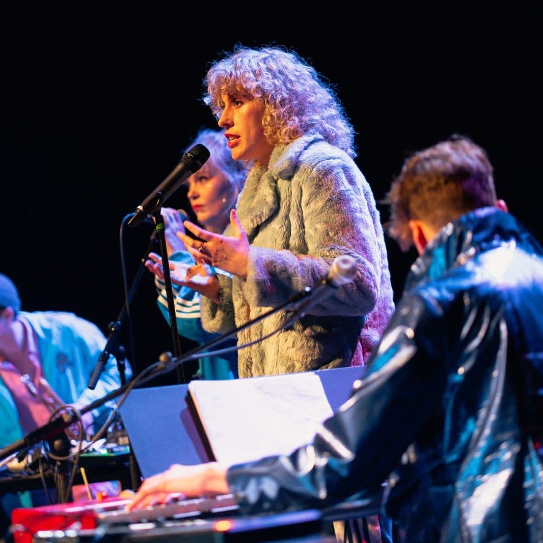 Vrouw staat bij microfoon op een podium met muzikanten om haar heen.